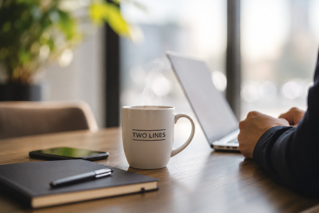 Two Lines-kopp med dampende kaffe på trebord, med laptop og notatbok